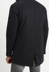 Parka negra con un cuello alto, detalles de botones y una abertura en la parte trasera. Fabricada de un tejido texturizado con un frontal oculto.