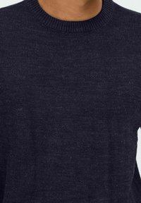 Nahaufnahme einer Person, die einen dunkelblauen gestrickten Pullover mit ribbelndem Rundhalsausschnitt trägt.