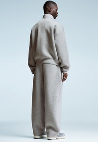 Uomo che indossa una felpa oversize grigia chiara e pantaloni larghi, in piedi di lato su uno sfondo chiaro.