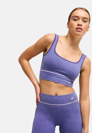 Young woman wearing a ribbed purple sports bra and high-waisted leggings with subtle pink stripes on a plain light background.