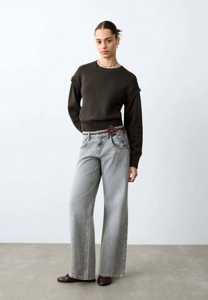 Donna in piedi che indossa un maglione marrone scuro, jeans a gamba larga grigio chiaro con una cintura color bordeaux e scarpe marroni su uno sfondo bianco uniforme.