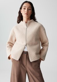 Giacca zip-up beige chiaro realizzata in tessuto testurizzato, con colletto alto e vestibilità rilassata. Abbinata a pantaloni brown a gamba larga e su misura.