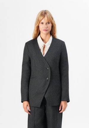 Mørk grå tettsittende blazer med omslagdesign, to knapper og sidelommer. Båret over en hvit bluse med rysjer ved kragen.