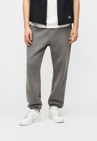 Άτομο που φορά γκρι φόρμα, λευκά αθλητικά παπούτσια, λευκό πουκάμισο και μαύρο γιλέκο, στέκεται μπροστά σε λευκό φόντο.