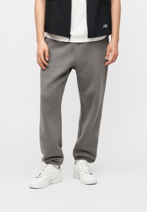 Άτομο που φορά γκρι φόρμα, λευκά αθλητικά παπούτσια, λευκό πουκάμισο και μαύρο γιλέκο, στέκεται μπροστά σε λευκό φόντο.