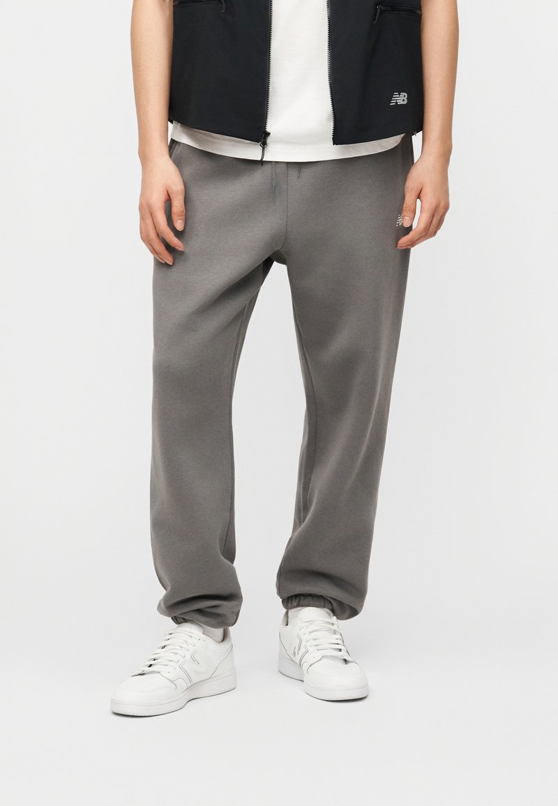 Άτομο που φορά γκρι φόρμα, λευκά αθλητικά παπούτσια, λευκό πουκάμισο και μαύρο γιλέκο, στέκεται μπροστά σε λευκό φόντο.