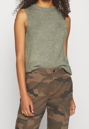 Kvinde iført ærmeløs olivengrøn strik top og brune camouflage cargobukser, med krøllet hår synligt ved skuldrene.