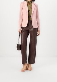 Světle růžový blazer přes zelený květinový top, zkombinovaný s hnědými kalhotami z umělé kůže a béžovými sandály na podpatku. Doplňkem je malá černá kabelka.