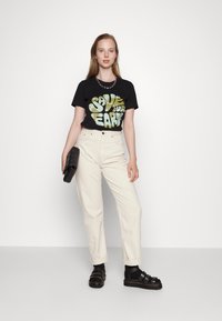Camisa de algodão preta com gráfico "SAVE OUR EARTH" em verde, combinada com jeans de cintura alta na cor creme e sandálias pretas. Bolsa preta.