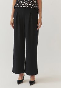 Zwarte wijde broek met plooien, gemaakt van een zachte stof. Heeft een gladde tailleband en wordt gedragen met zwarte puntige hakken.