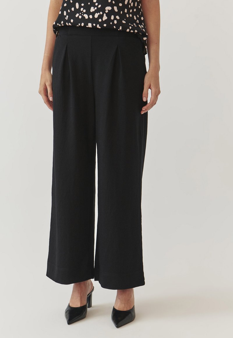 Zwarte wijde broek met plooien, gemaakt van een zachte stof. Heeft een gladde tailleband en wordt gedragen met zwarte puntige hakken.