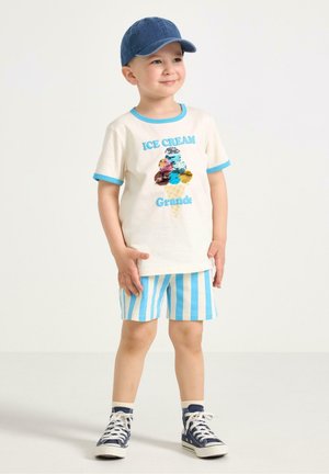 Ung dreng iført blå kasket, hvid T-shirt med is-grafik og tekst, blåstribede shorts, mørkeblå sneakers, står med hænderne ned langs siderne.