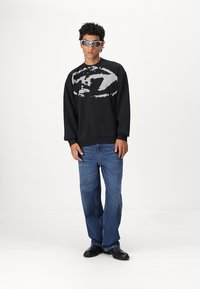 Sort sweatshirt med et stort gråt grafisk design, parret med blå vide jeans og sorte sko. Model iført reflekterende solbriller.