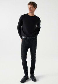 Suéter negro de cuello redondo, jeans negros ajustados y zapatos negros con cordones. La tela es lisa y de color sólido. Cinturón estándar incluido.