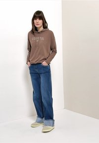 Femme debout, mains dans les poches, portant un sweat à capuche brun « Coastal », un jean bleu large avec des bords retroussés et des baskets beige, devant des murs blancs.