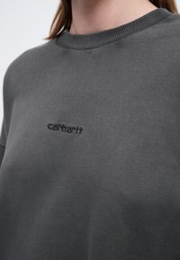 Sudadera de color gris oscuro hecha de un tejido texturizado, con cuello redondo y un sutil logo bordado en el pecho. Diseño sencillo.