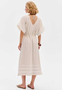 Beige jurk met een V-rug, korte brede mouwen en een koord in de taille. Kenmerkt zich door witte zigzagaccenten langs de zoom. Textuurstof.