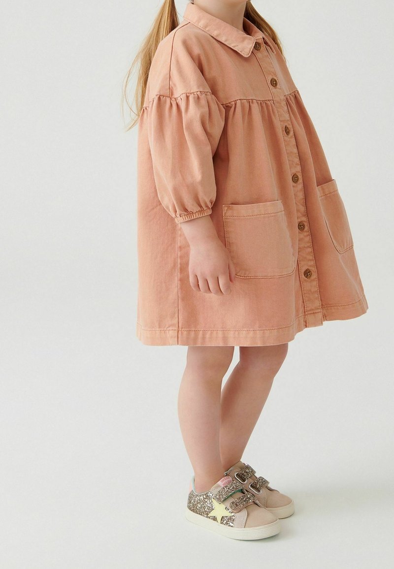 Enfant portant une robe pêche boutonnée avec des manches ballon et des poches devant, debout et portant des baskets beige avec paillettes dorées et motif d'étoile.