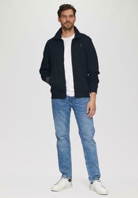 Veste zippée bleu foncé en matériau doux, portée sur une chemise blanche, associée à un jean bleu clair et des baskets blanches.