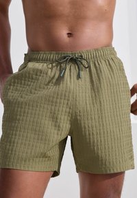 Olivgrüne Badeshorts aus strukturiertem Stoff, mit elastischem Bund und Kordelzugverschluss. Sie verfügen über seitliche Taschen und eine lockere Passform.