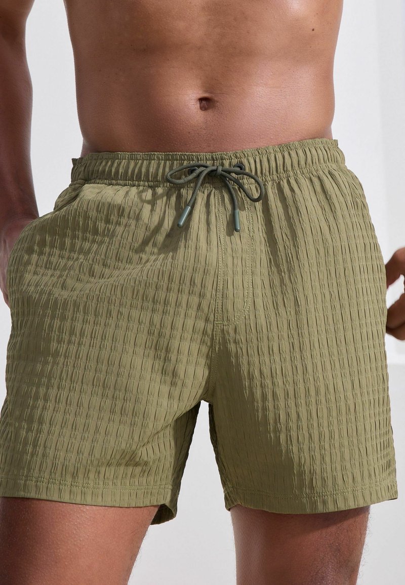 Olivgrüne Badeshorts aus strukturiertem Stoff, mit elastischem Bund und Kordelzugverschluss. Sie verfügen über seitliche Taschen und eine lockere Passform.
