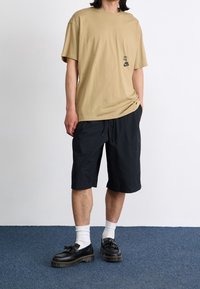 Camiseta de algodón beige con logo negro, combinada con pantalones cortos negros, calcetines blancos y zapatos negros de cordones con suela gruesa.
