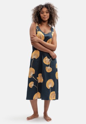 Woman with curly hair stands barefoot wearing a sleeveless navy dress with large yellow leaf patterns on a white background.