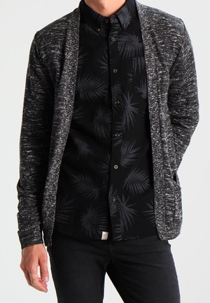 Mand iført en sort skjorte med knapper og mørke palmebladmønstre under en tekstureret grå cardigan med åbent front og sorte bukser.