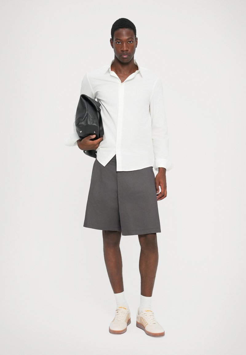 Witte overhemd met lange mouwen, grijze shorts en beige sneakers met oranje accenten. Zwarte tas over de linkerschouder. Glad materiaal en op maat gemaakte pasvorm.