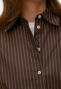 Brązowa koszula zapinana na guziki z białymi pionowymi paskami, posiadająca klasyczny kołnierzyk i dwa białe guziki przy dekolcie.