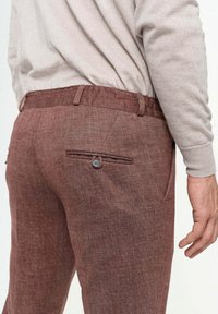 Pantalones marrones con textura y un sutil patrón de espiga, que cuentan con un bolsillo trasero con botón y presillas para cinturón. Tela arrugada y ajuste entallado.