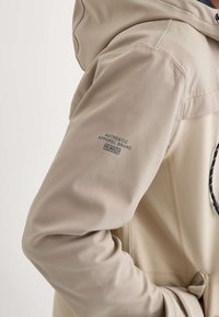 Veste beige avec capuche, affichant un gros plan du bras gauche avec le texte de marque "AUTHENTIC APPAREL BRAND MÜNICH."