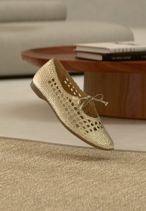 Chaussure plate tissée couleur or avec un design perforé et un détail de nœud, dotée d'une semelle en caoutchouc. Présentée sur un tapis texturé et un mobilier minimaliste.