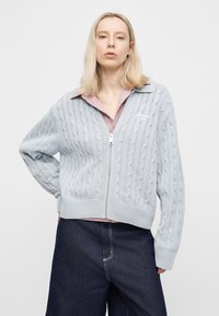 Ξανθιά γυναίκα που φοράει ένα ανοιχτό γαλάζιο πουλόβερ με φερμουάρ και πλεκτό σχέδιο σε σχήμα καλωδίου, πάνω από ένα ροζ πουκάμισο με γιακά και σκούρο μπλε παντελόνι ψηλόμεσο, στέκεται με το ένα χέρι στη μέση.