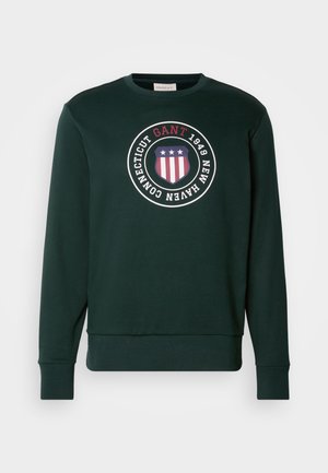 Ciemnozielona bluza z okrągłym dekoltem, długimi rękawami i okrągłym emblematem z napisem "GANT 1949 New Haven Connecticut" z tarczą w gwiazdy i paski.