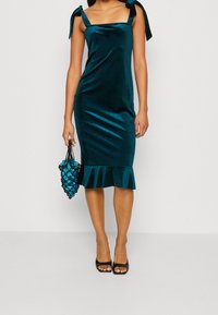 Teal sammetklänning med fyrkantig halsringning och knytremmar, med en volangkant. Modellen håller en blå handväska och bär svarta öppna klackar.