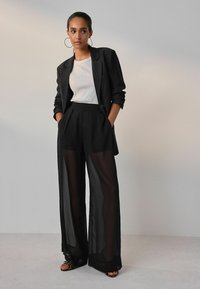 Black transparent wide-leg pants with pleats and a matching oversized blazer over a white ribbed t-shirt. Hoop earrings complete the look.