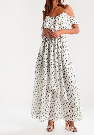 Kvinde iført en hvid maxi-kjole med bare skuldre og små sorte blomsterprint samt beige højhælede sandaler med sløjfer.