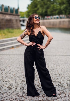 Žena v černém volném kombinézovém oblečení a slunečních brýlích stojí na dlažebních kostkách vedle vody, usmívá se s rukama na bocích a ukazuje tetování na pažích.