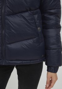 Giacca a vento blu navy con sezioni trapuntate, chiusura con zip e dettagli in metallo a scatto. Presenta polsini elasticizzati per calore e comfort.