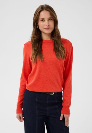 Femme aux longs cheveux bruns portant un pull orange à manches longues et un jean bleu foncé sur un fond clair uni.