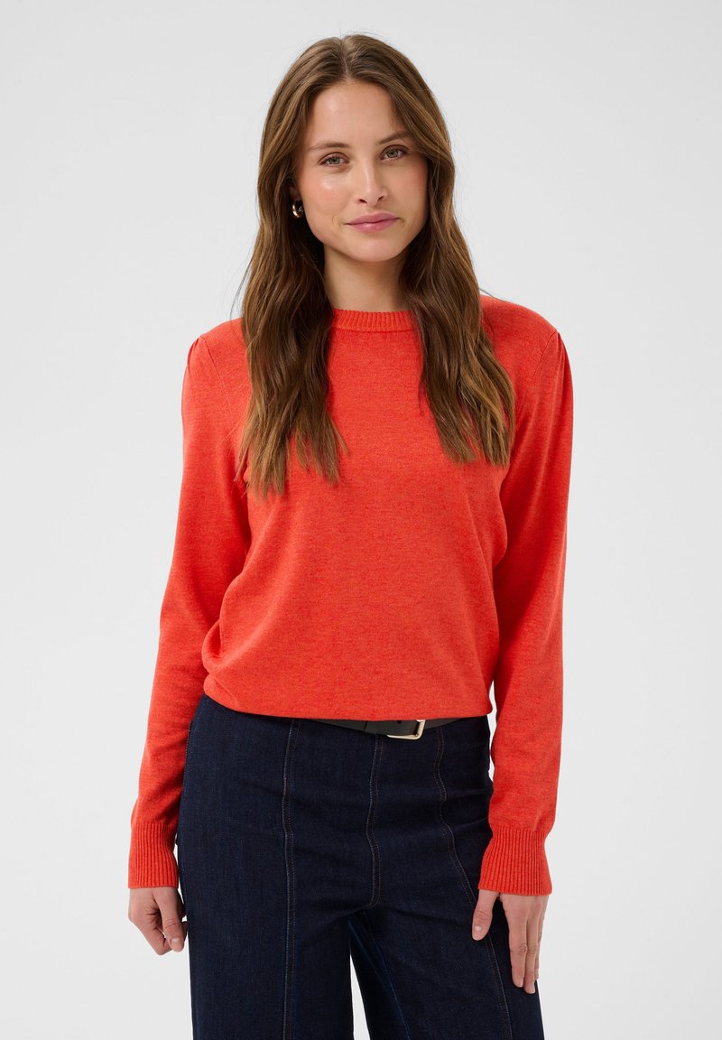 Femme aux longs cheveux bruns portant un pull orange à manches longues et un jean bleu foncé sur un fond clair uni.