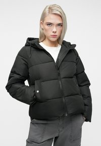 Jauna sieviete ar blondiem matiem, tērpta melnā pūkainājā jakā ar kapuci, baltā kreklā un pelēkās biksēs, stāv ar rokām kabatās.