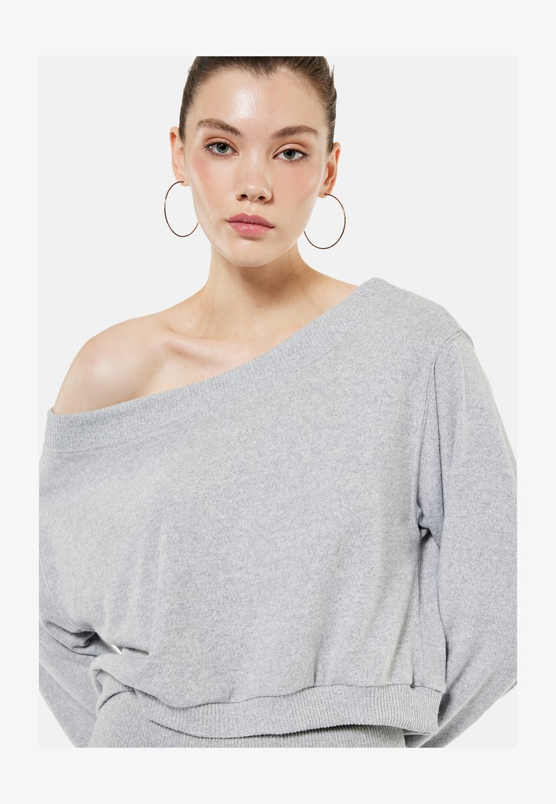 Young woman with light skin and green eyes wears large hoop earrings and a gray off-shoulder sweater against a plain background.