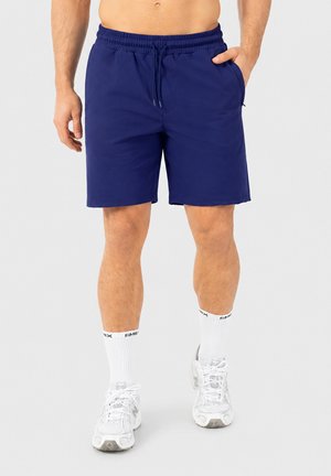 Homme portant un short bleu marine à cordon, des chaussettes blanches montantes et des chaussures de sport blanches, debout devant un fond clair uni.