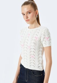 Hvid strikket sweater med et struktureret mønster og lyserøde sløjfe detaljer. Korte ærmer og en rund hals fuldender designet.