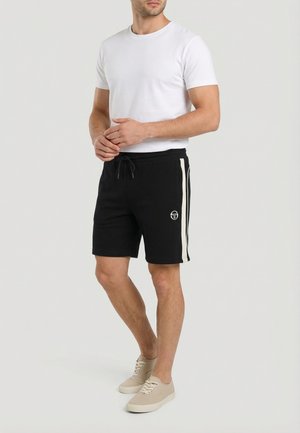 Uomo con camicia bianca a maniche corte, pantaloncini neri con coulisse e strisce laterali bianche, e sneaker beige con lacci, in piedi con le mani giunte.