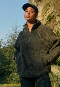 Olijfgroene fleece pullover met een hoge kraag, rits tot een kwart en twee voorzakken. Heeft een gestructureerde stof en contrasterende naden.
