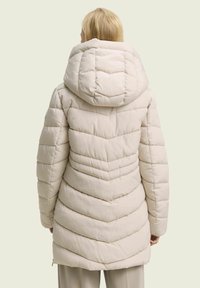Manteau rembourré de couleur beige clair avec une grande capuche, présentant un motif matelassé en chevrons, des manches longues et une coupe mi-cuisse.