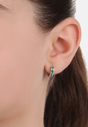 Silberne Creolen mit grünen Edelsteinen, die ein elegantes, geschwungenes Design und eine glatte Oberfläche aufweisen, werden an einem Ohr präsentiert.